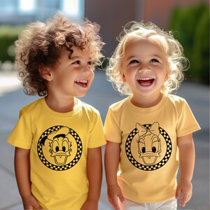 May include: Two children wearing yellow t-shirts with black and white cartoon duck designs. The girl's shirt features a duck with a bow, and the boy's shirt features a duck wearing a cap.
