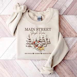 May include: Beige sweatshirt with the text "Main Street Sleigh Rides" and a graphic of Mickey and Minnie Mouse sledding. The design includes pine trees and the phrases "Hot Cocoa," "Cookies," and "Magical Fun."