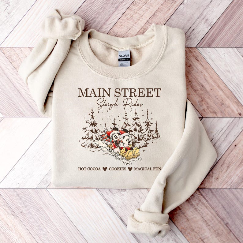 May include: Beige sweatshirt with the text "Main Street Sleigh Rides" and a graphic of Mickey and Minnie Mouse sledding. The design includes pine trees and the phrases "Hot Cocoa," "Cookies," and "Magical Fun."