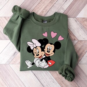 May include: A green sweatshirt with a graphic of Mickey Mouse and Minnie Mouse hugging. Three pink hearts are above the characters.