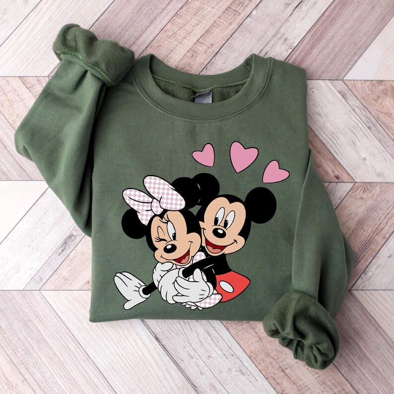 May include: A green sweatshirt with a graphic of Mickey Mouse and Minnie Mouse hugging. Three pink hearts are above the characters.