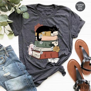 May include: A heather dark grey t-shirt with a black cat sleeping on a stack of books. The books are decorated with flowers and have a vintage look.