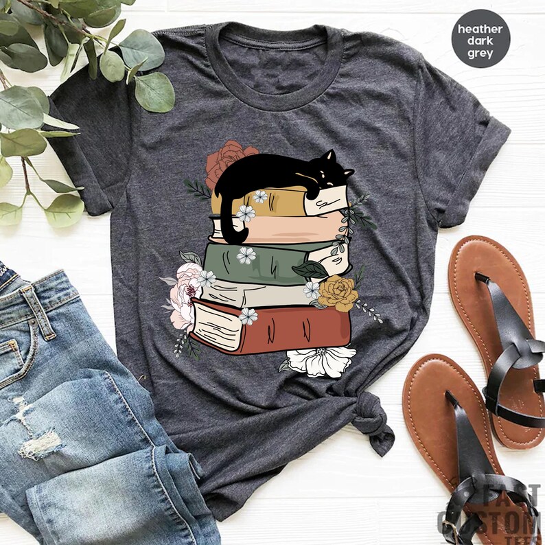 May include: A heather dark grey t-shirt with a black cat sleeping on a stack of books. The books are decorated with flowers and have a vintage look.