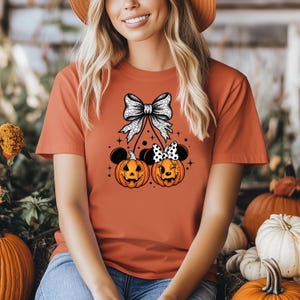May include: Orange t-shirt featuring a Halloween design. The design includes two jack-o'-lanterns with mouse ears and bows, connected to a large bow at the top. The pumpkins have smiling faces. The shirt is a solid orange color.