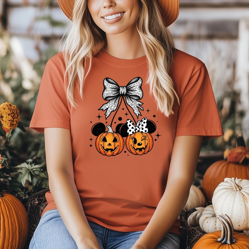 May include: Orange t-shirt featuring a Halloween design. The design includes two jack-o'-lanterns with mouse ears and bows, connected to a large bow at the top. The pumpkins have smiling faces. The shirt is a solid orange color.
