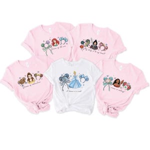 May include: A collection of pink and white t-shirts featuring Disney princess designs. The shirts showcase Ariel, Belle, Cinderella, Mulan, and Moana. Each tee has a unique graphic with phrases such as "Dream of the sea".