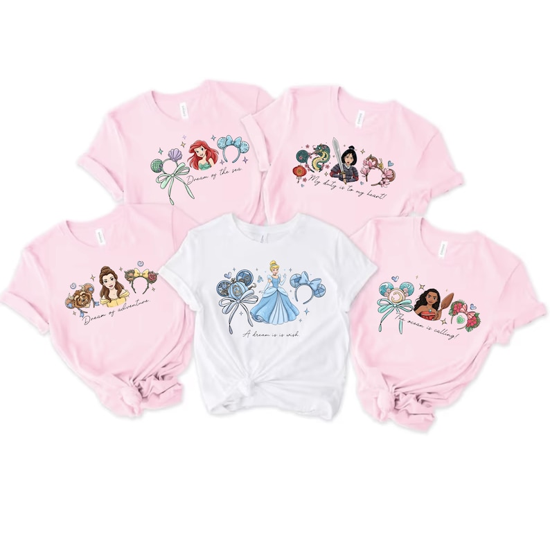 May include: A collection of pink and white t-shirts featuring Disney princess designs. The shirts showcase Ariel, Belle, Cinderella, Mulan, and Moana. Each tee has a unique graphic with phrases such as "Dream of the sea".