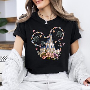 May include: Black t-shirt featuring a colorful castle design with floral accents and fireworks. The castle is surrounded by a Mickey Mouse-shaped design made of flowers and Mickey Mouse heads. The shirt is a classic crew neck.