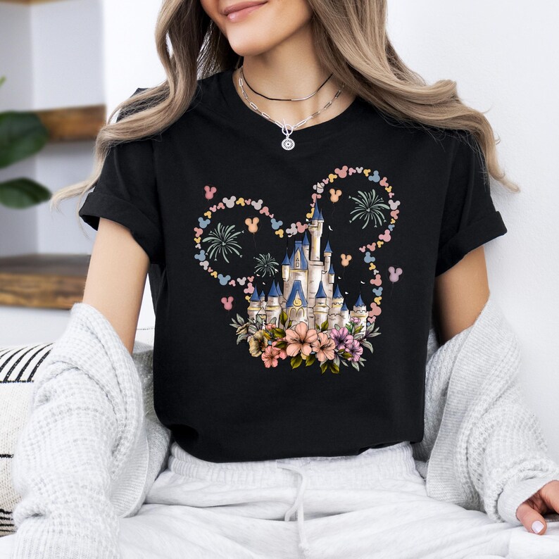 May include: Black t-shirt featuring a colorful castle design with floral accents and fireworks. The castle is surrounded by a Mickey Mouse-shaped design made of flowers and Mickey Mouse heads. The shirt is a classic crew neck.