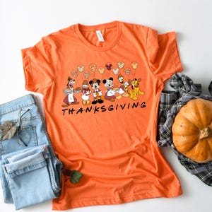K&ouml;nnte beinhalten: Orangefarbenes T-Shirt mit Thanksgiving-Motiv mit Mickey Mouse und seinen Freunden. Das Shirt hat ein Motiv mit Mickey Mouse, Minnie Mouse, Donald Duck, Goofy, Pluto und Chip und Dale. Das Motiv enth&auml;lt den Text "THANKSGIVING".