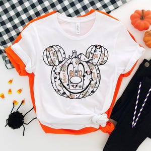 May include: White t-shirt featuring a black and white graphic of a pumpkin Mickey Mouse head. The pumpkin is decorated with Halloween-themed patterns and text, including "Spooky" and "Boo".
