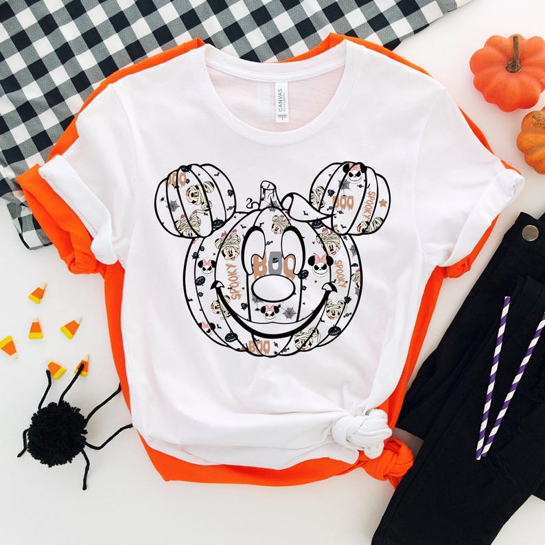 May include: White t-shirt featuring a black and white graphic of a pumpkin Mickey Mouse head. The pumpkin is decorated with Halloween-themed patterns and text, including "Spooky" and "Boo".