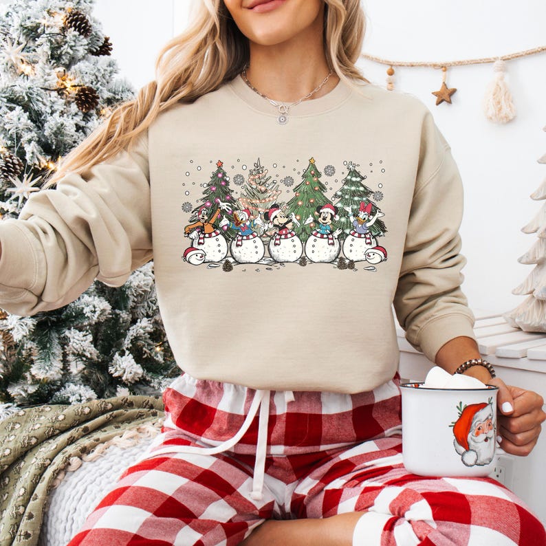 Op de afbeelding: Een beige sweatshirt met een kerstthema ontwerp met Mickey Mouse en zijn vrienden verkleed als sneeuwpoppen. Het ontwerp bevat vijf sneeuwpoppen, elk met een ander personage, die voor vijf kerstbomen staan. De sneeuwpoppen dragen hoeden en sjaals, en de bomen zijn versierd met ornamenten en lampjes. Het sweatshirt heeft lange mouwen en een ronde hals.