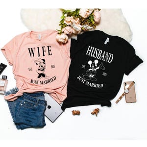 May include: Two t-shirts, one pink and one black, with the words "Wife" and "Husband" printed on them. Each shirt features a cartoon image of Mickey Mouse or Minnie Mouse and the text "Est. 2024 Just Married".