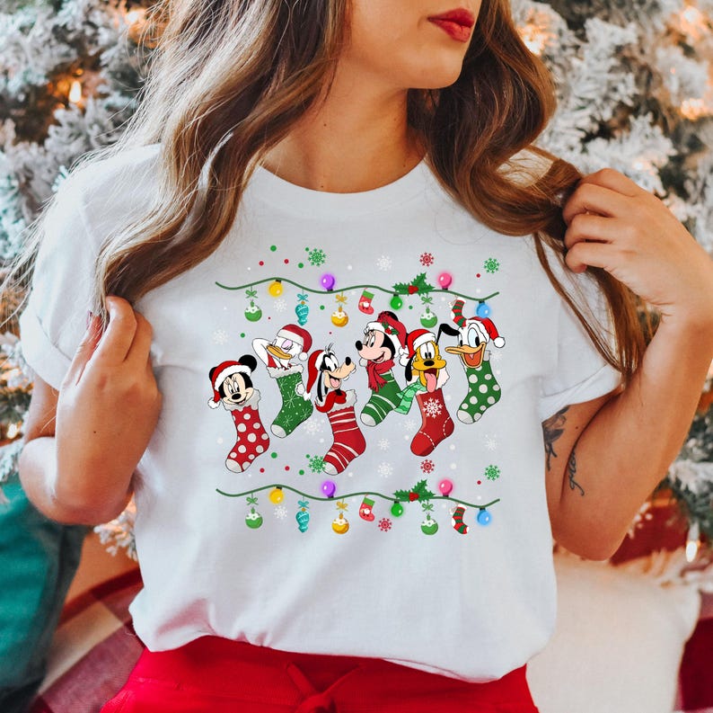 May include: White t-shirt with a Christmas design featuring Mickey Mouse, Minnie Mouse, Donald Duck, Goofy, and Pluto wearing Christmas stockings. The design includes a string of Christmas lights and ornaments.