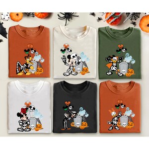 May include: Six Halloween-themed t-shirts in various colors, each featuring a cartoon character in a skeleton costume. The designs include Mickey Mouse, Minnie Mouse, Donald Duck, and Pluto, with pumpkins and tombstones. The shirts are orange, cream, green, white, black, and rust.