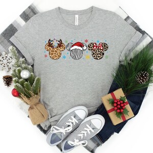 May include: A gray t-shirt with a Christmas-themed design featuring three animal-themed Mickey Mouse ears. The ears are decorated with a reindeer, a zebra, and a leopard print. The shirt is a great gift for Disney fans.