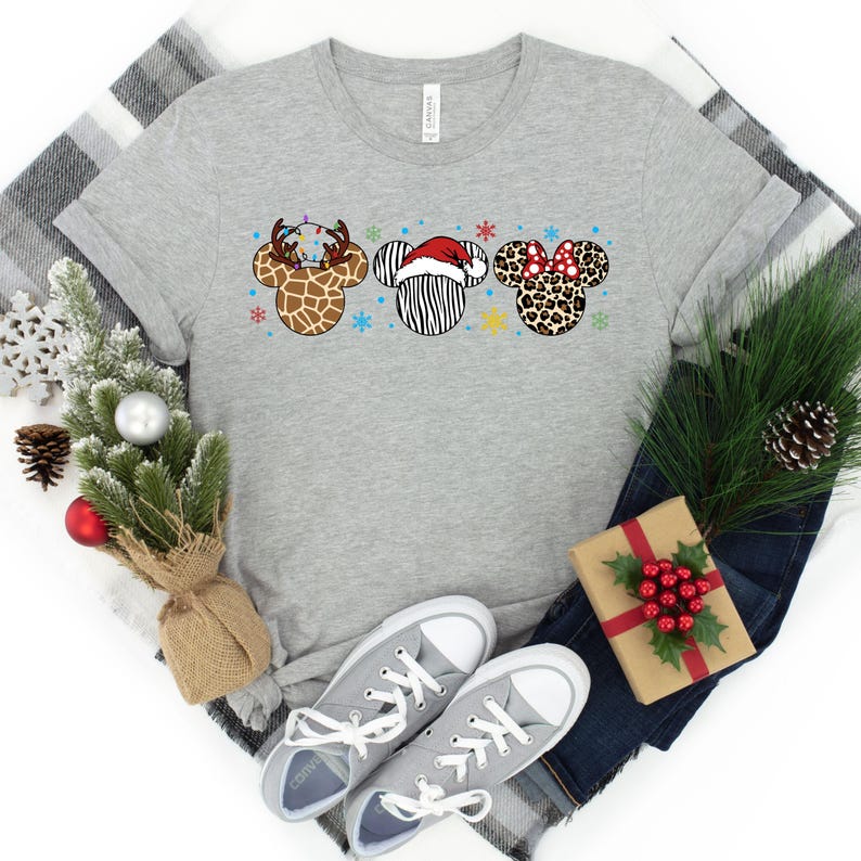 May include: A gray t-shirt with a Christmas-themed design featuring three animal-themed Mickey Mouse ears. The ears are decorated with a reindeer, a zebra, and a leopard print. The shirt is a great gift for Disney fans.