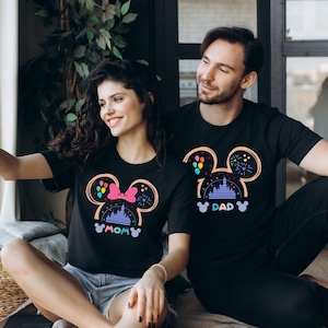 May include: A couple wearing matching black t-shirts with a Mickey Mouse silhouette design. The woman's shirt says "MOM" and the man's shirt says "DAD". The design features a pink bow, a castle, balloons, and fireworks.
