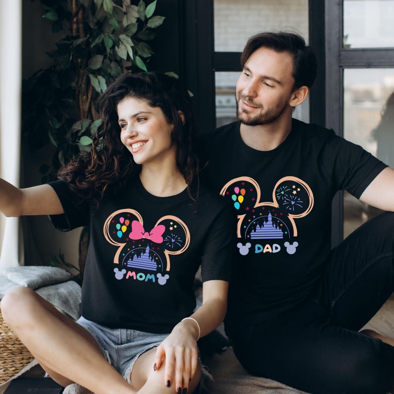 May include: A couple wearing matching black t-shirts with a Mickey Mouse silhouette design. The woman's shirt says "MOM" and the man's shirt says "DAD". The design features a pink bow, a castle, balloons, and fireworks.
