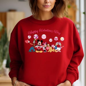 May include: Red sweatshirt with a graphic of Mickey Mouse, Minnie Mouse, Goofy, Donald Duck, and Pluto holding hearts and balloons. The text "Happy Valentine's Day" is above the characters.