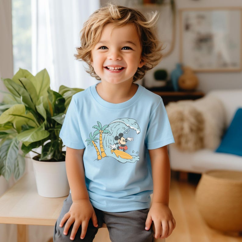 May include: A light blue t-shirt featuring a graphic of Mickey Mouse surfing on a wave with palm trees in the background.