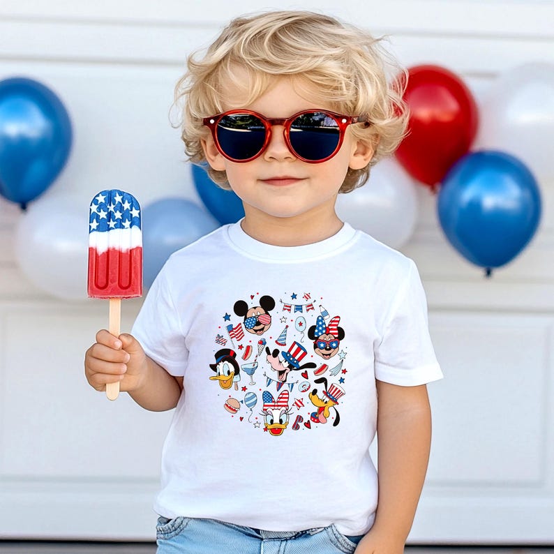 May include: A young child wearing red sunglasses and a white t-shirt with a colorful graphic of Disney characters in patriotic attire. The child is holding a red, white, and blue popsicle. Balloons in red, white, and blue are in the background.