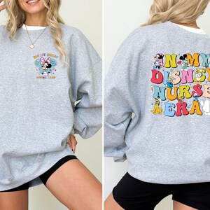 May include: A light gray sweatshirt with a colorful graphic on the front and back. The front features Minnie Mouse with text, and the back displays the words "In My Disney Nurse Era" with Disney characters. The sweatshirt is a casual style.