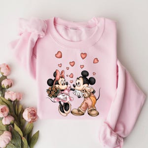May include: Pink sweatshirt with a graphic of Mickey Mouse and Minnie Mouse holding hands with hearts around them.