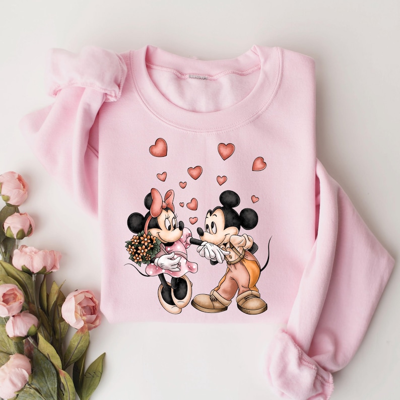May include: Pink sweatshirt with a graphic of Mickey Mouse and Minnie Mouse holding hands with hearts around them.