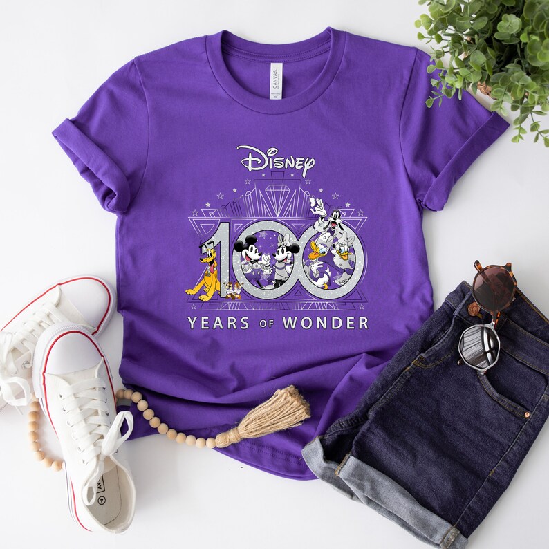 May include: Purple t-shirt with a graphic of Mickey Mouse and friends celebrating Disney's 100th anniversary. The graphic includes the text "Disney 100 Years of Wonder".