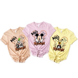 Puede incluir: Tres camisetas con tem&aacute;tica de Halloween con Mickey Mouse y Goofy. Las camisetas son de color beige, rosa y amarillo. Cada camiseta tiene una imagen de dibujos animados de Mickey Mouse o Goofy con el texto "BOO!" y "HALLOWEEN".