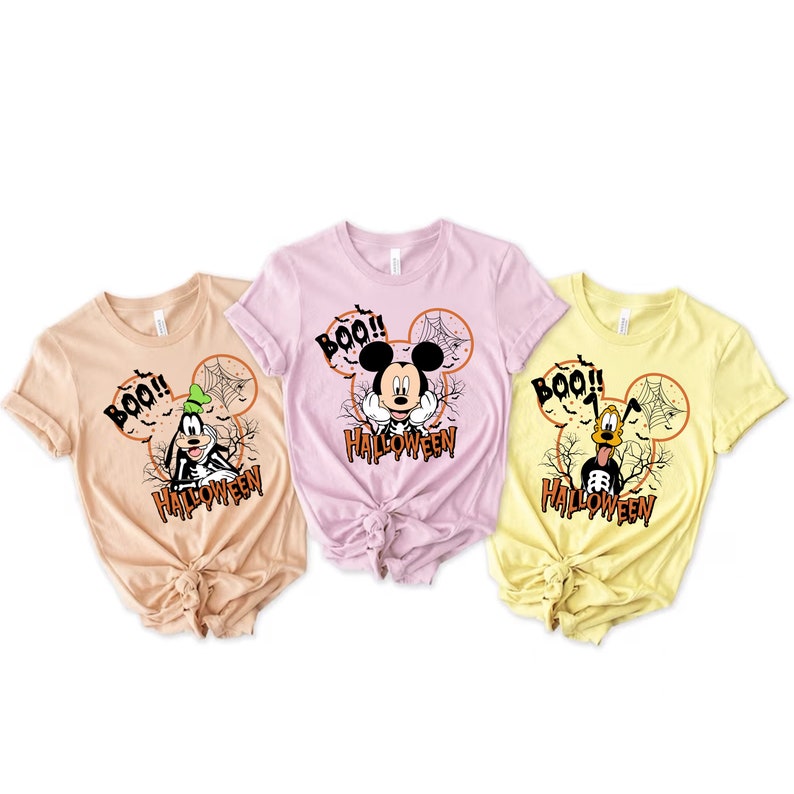 Puede incluir: Tres camisetas con tem&aacute;tica de Halloween con Mickey Mouse y Goofy. Las camisetas son de color beige, rosa y amarillo. Cada camiseta tiene una imagen de dibujos animados de Mickey Mouse o Goofy con el texto "BOO!" y "HALLOWEEN".