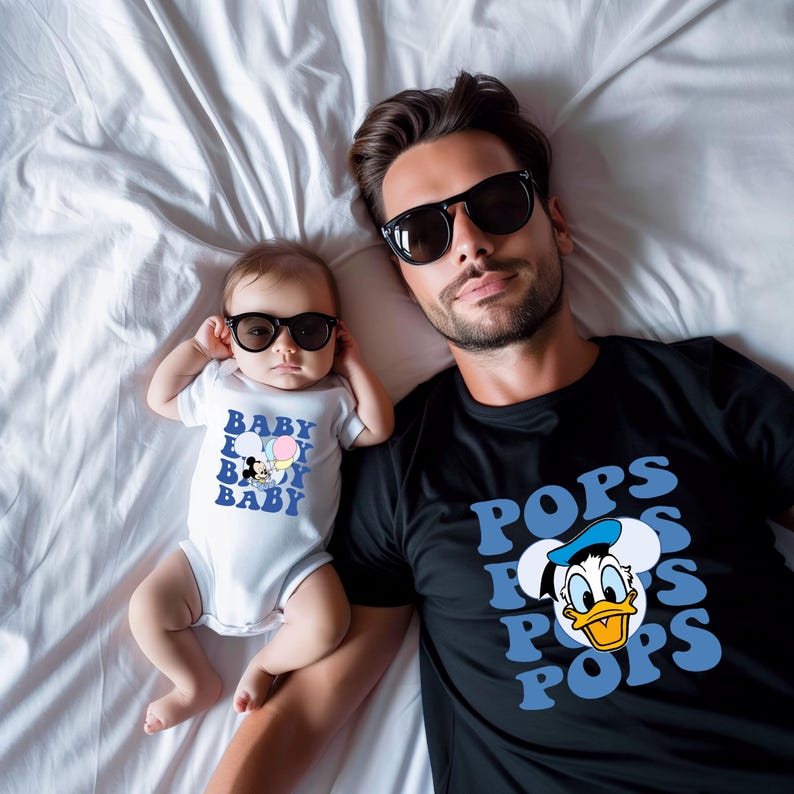 Puede incluir: Un padre y un beb&eacute; con gafas de sol, tumbados en una s&aacute;bana blanca. El beb&eacute; lleva un body blanco con un dibujo de Mickey Mouse y el texto "BABY BOY BABY". El padre lleva una camiseta negra con un dibujo de Pato Donald y el texto "POPS".