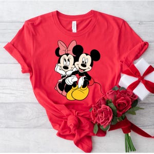 May include: Red t-shirt with a graphic of Mickey Mouse and Minnie Mouse holding hands.