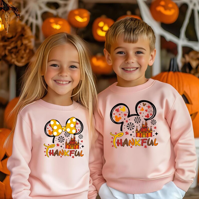 Puede incluir: Dos ni&ntilde;os con sudaderas rosas a juego con un dise&ntilde;o de orejas de Mickey Mouse. El dise&ntilde;o incluye un lazo amarillo, hojas de oto&ntilde;o, un castillo y la palabra "THANKFUL". Calabazas y decoraci&oacute;n oto&ntilde;al en el fondo.
