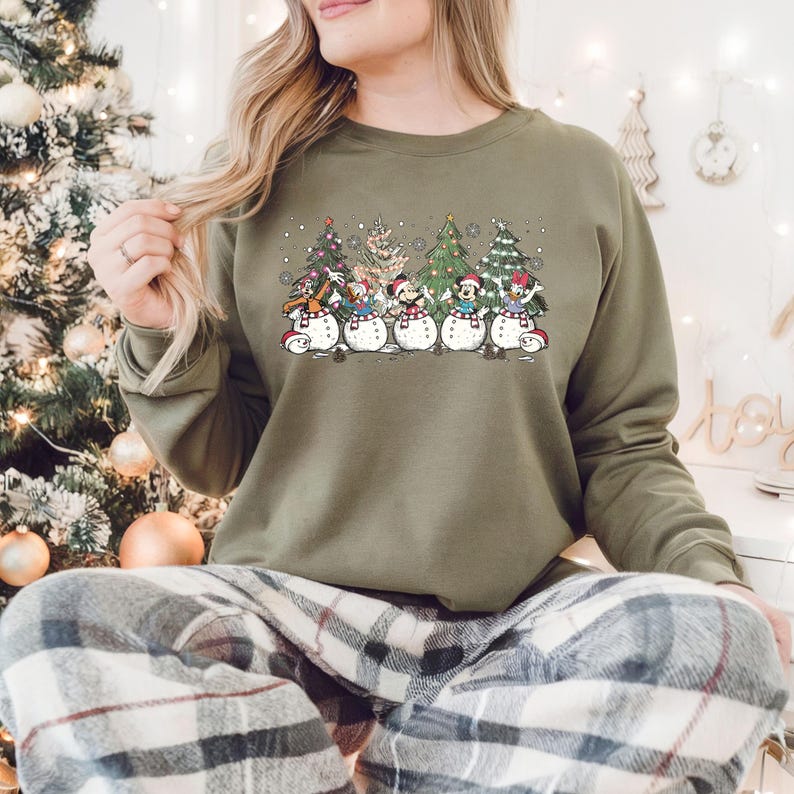 Op de afbeelding: Een groen sweatshirt met een kerstthema ontwerp met cartoonfiguren die voor sneeuwpoppen staan. De figuren dragen kerstmanmutsen en houden cadeautjes vast. Het ontwerp is omringd door sneeuwvlokken en kerstboom.