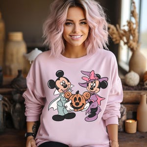 May include: A pink sweatshirt with a graphic print of Mickey Mouse and Minnie Mouse dressed in Halloween costumes. Mickey is dressed as a vampire and Minnie is dressed as a witch. They are holding a jack-o'-lantern.