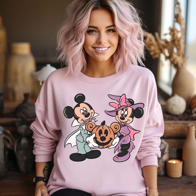 May include: A pink sweatshirt with a graphic print of Mickey Mouse and Minnie Mouse dressed in Halloween costumes. Mickey is dressed as a vampire and Minnie is dressed as a witch. They are holding a jack-o'-lantern.