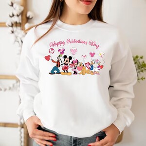 May include: White sweatshirt with a pink and white graphic of Mickey Mouse, Minnie Mouse, Goofy, Pluto, and Donald Duck holding heart-shaped balloons. The text "Happy Valentine's Day" is printed above the characters.