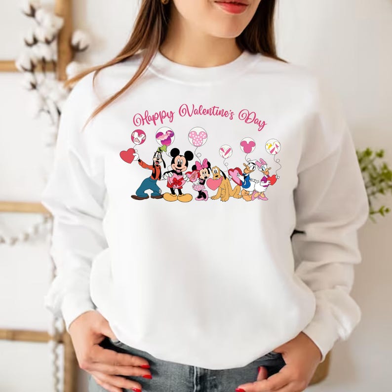 May include: White sweatshirt with a pink and white graphic of Mickey Mouse, Minnie Mouse, Goofy, Pluto, and Donald Duck holding heart-shaped balloons. The text "Happy Valentine's Day" is printed above the characters.