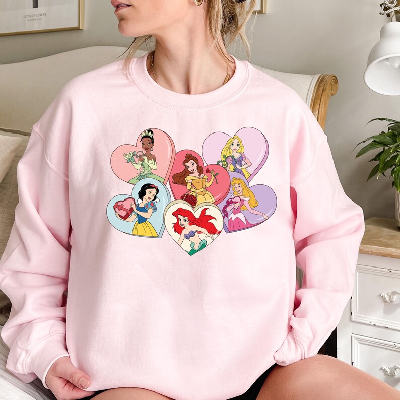 Pode incluir: Camisola rosa com um gr&aacute;fico de princesas Disney em cora&ccedil;&otilde;es. As princesas incluem Branca de Neve, Cinderela, Ariel, Bela, Aurora e Tiana.