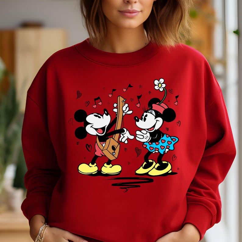 Pode incluir: Camisola vermelha com um gr&aacute;fico de Mickey Mouse e Minnie Mouse tocando m&uacute;sica. Mickey est&aacute; tocando uma guitarra e Minnie est&aacute; segurando uma flor.