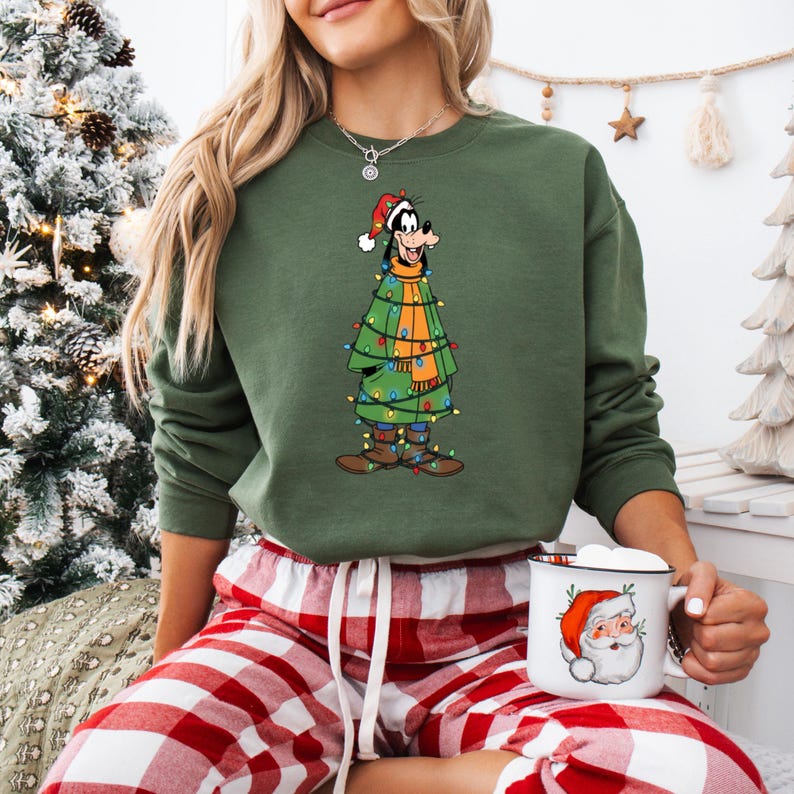 May include: Green sweatshirt featuring a cartoon character dressed as a Christmas tree, adorned with colorful lights and a Santa hat. The character is wearing an orange scarf and brown boots. The person is wearing red and white plaid pajama pants and holding a mug with a Santa Claus design.