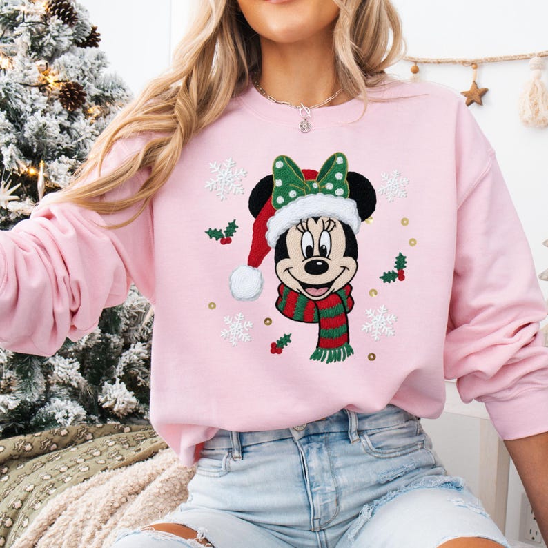 May include: A light pink sweatshirt featuring a Minnie Mouse graphic in a Santa hat, green bow, and striped scarf. The design includes snowflakes and holly accents, perfect for the holiday season.
