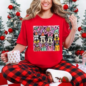 Op de afbeelding: Een rood T-shirt met een kleurrijke afbeelding van Disney-figuren in kerstkleding. De personages zijn Goofy, Mickey, Minnie en Donald Duck. De persoon draagt een rood-zwarte geruite pyjamabroek en houdt een mok vast.