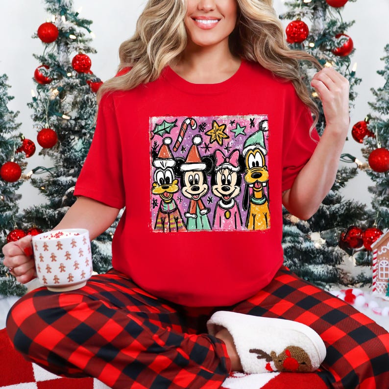 Op de afbeelding: Een rood T-shirt met een kleurrijke afbeelding van Disney-figuren in kerstkleding. De personages zijn Goofy, Mickey, Minnie en Donald Duck. De persoon draagt een rood-zwarte geruite pyjamabroek en houdt een mok vast.