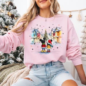 Op de afbeelding: Een lichtroze sweatshirt met een winterse sc&egrave;ne met Mickey en Minnie Mouse. De afbeelding toont het paar voor een kerstboom en kleurrijke gebouwen. De sweatshirt is een crew neck stijl.