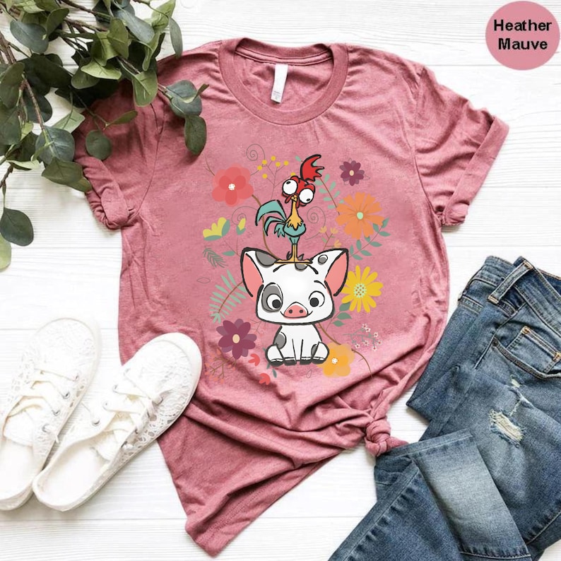 Puede incluir: Una camiseta de color malva jaspeado con una imagen de dibujos animados de un cerdo y un gallo en un dise&ntilde;o floral. La camiseta se muestra sobre una superficie blanca con un par de zapatillas blancas y unos vaqueros azules.