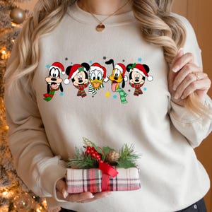 May include: A beige sweatshirt with a festive design featuring Mickey Mouse, Minnie Mouse, Goofy, Pluto, and Donald Duck wearing Santa hats. The characters are lined up across the front of the sweatshirt.